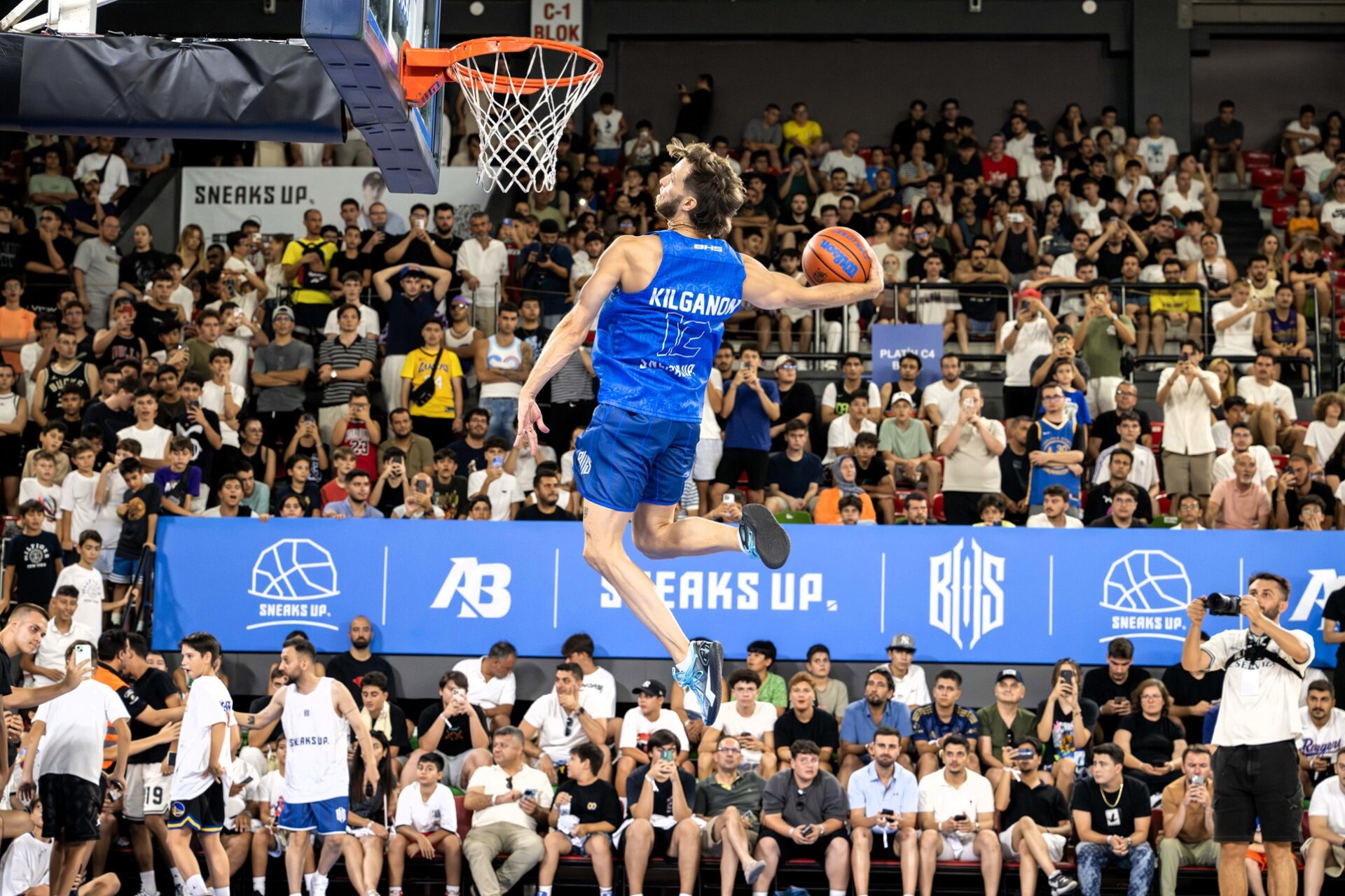 İzmir, bu yazın en enerjik ve en coşkulu basketbol etkinliğine ev sahipliği yaptı. Sneaks Up sponsorluğunda düzenlenen Brotherhood Squad (BHS) Smaç Gösterisi, Mustafa Kemal Atatürk Karşıyaka Spor Salonu’nda üçüncü kez gerçekleştirildi. Binlerce basketbolseverin tribünleri doldurduğu etkinlik, unutulmaz anlara sahne oldu. Jordan Kilganon İzmir’i Ayağa Kaldırdı Dünyaca ünlü Kanadalı dunker Jordan Kilganon, akrobatik smaçlarıyla geceye damga vurdu. Tribünlerdeki heyecanı zirveye taşıyan performansı, sadece basketbol değil aynı zamanda bir gösteri sanatına dönüştü. Smaç şovu boyunca izleyiciler nefesini tuttu, her sıçrayış bir sonraki alkış tufanının habercisi oldu. Sadece Smaç Değil, Tam Bir Spor ve Eğlence Şöleni BHS Smaç Gösterisi yalnızca smaçlarla sınırlı değildi. Etkinlikte üçlük yarışmaları, birebir kapışmalar, dans gösterileri ve ünlü isimlerin katıldığı yetenek yarışmaları yer aldı. Gecenin organizasyonunu üstlenen içerik üreticisi Alper Biçen ve BHS ekibi, her detayıyla izleyenlere unutulmaz bir deneyim sundu. Khontkar’dan BHS’ye Özel Şarkı Türkçe hiphop sahnesinin önemli isimlerinden Khontkar, BHS etkinliğine özel olarak hazırladığı şarkısıyla geceye ayrı bir hava kattı. Dinamik sahne performansı ve özel prodüksiyonuyla atmosferi bambaşka bir seviyeye taşıyan Khontkar, etkinliğin enerjisini doruk noktaya ulaştırdı. Ünlü Jüri Heyeti Gecenin Nabzını Tuttu Jüri koltuğunda tanıdık isimler vardı: Spor yorumcusu Kaan Kural Amerikalı profesyonel basketbolcu Jordan Theodore Gazeteci Deniz Aksoy Fenerbahçe basketbolcusu Tarık Biberovic Jüri heyeti, gece boyunca düzenlenen All Star maçı, üçlük yarışmaları ve yetenek kapışmalarını değerlendirdi. Tarık Biberovic’in de sahaya çıkarak üçlük yarışmasında performans sergilemesi geceye renk kattı. Memo ve Med Brodie Kapışmaları Nefes Kesti Etkinliğin sürprizlerinden biri, geçen yılın şampiyonu Med Brodie (Muhammed Sanyang) ile Amerikalı sokak basketbolu fenomeni Moon’un birebir kapışmasıydı. Kazanan Med oldu. Ayrıca Ankara’dan çıkan ve kariyerine ABD’de devam eden Memo (Mehmet Bener) ile 2018 Hillsborough All Star Şampiyonu Slim (Isaiah Hodge) karşı karşıya geldi. Kıran kırana geçen mücadelede kazanan Memo oldu. Sadece İzlemekle Kalmadılar: Katılımcılar da Kazandı BHS Smaç Gösterisi, izleyicileri sadece izleyici olarak değil, etkinliğin parçası olarak da sahaya çekti. Orta sahadan şut yarışmasına katılan izleyicilerden isabet bulanlar, Bilkom sponsorluğunda PlayStation 5, kulaklık ve çeşitli aksesuarlarla ödüllendirildi. 🏀 Sonuç: BHS Smaç Gösterisi, İzmir’de Gerçek Bir Basketbol Festivali Yarattı Spor, müzik, eğlence ve coşkunun birleştiği bu etkinlik, sadece basketbolseverleri değil, tüm şehir halkını etkisi altına aldı. BHS Smaç Gösterisi, bir kez daha neden yılın en çok beklenen etkinliklerinden biri olduğunu kanıtladı.